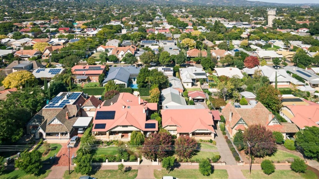 The image shows an aerial view of a serene suburban neighbourhood in Adelaide, illustrating a diverse Adelaide property portfolio. The scene captures a mix of residential homes with varying architectural styles and manicured gardens, some equipped with solar panels. The area is lush with mature trees and landscaping, highlighting the suburban charm and liveability ideal for property investments.