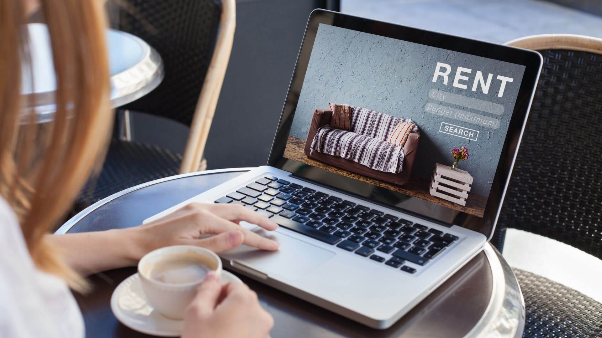 A woman is browsing rental property listings on a laptop at an outdoor cafe, the screen displaying a search interface with options for city and budget.