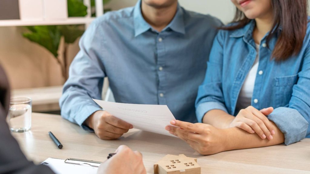 A couple is consulting a professional about selling their house, sitting at a table with documents spread out in front of them, emphasizing the careful consideration and planning involved in the home-selling process.