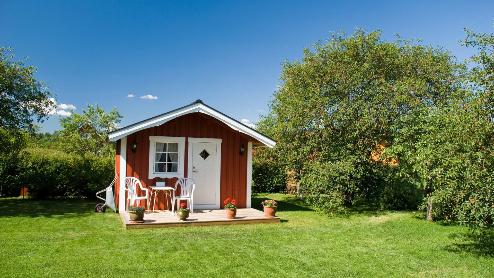 A charming red granny flat with a white door and windows, set in a lush green backyard surrounded by trees, providing a cozy and private space for additional living.