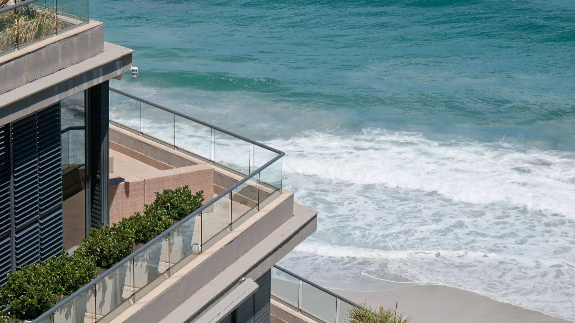 A modern seaside property overlooking the ocean, showcasing sleek architecture with a balcony surrounded by glass railings and lush greenery. The view highlights the importance of preparing your property for summer, ensuring it remains in prime condition for the warm months ahead.