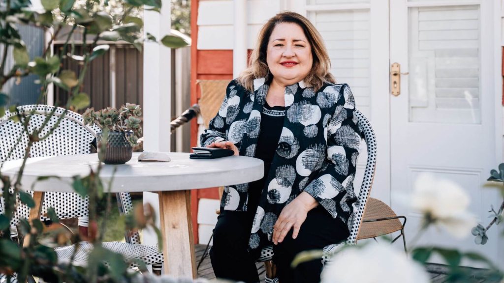 Amal, the Managing Director and a leading property manager in Adelaide, sitting at an outdoor table with a smile, surrounded by a cozy garden setting.