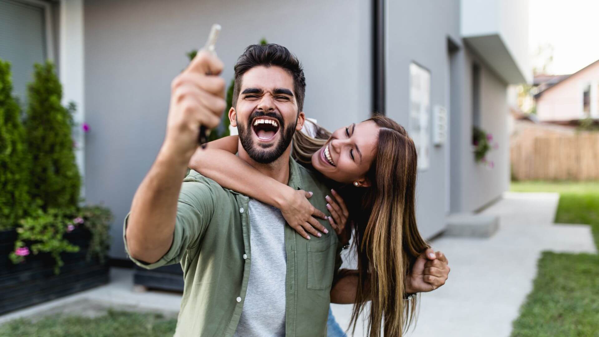 A joyful couple celebrating after successfully completing the purchase of a house. The man is holding a key up in the air with a big smile, while the woman hugs him from behind, both clearly excited about their new home. The modern house with a well-maintained garden can be seen in the background, symbolizing a fresh start and new beginnings in their journey as homeowners.