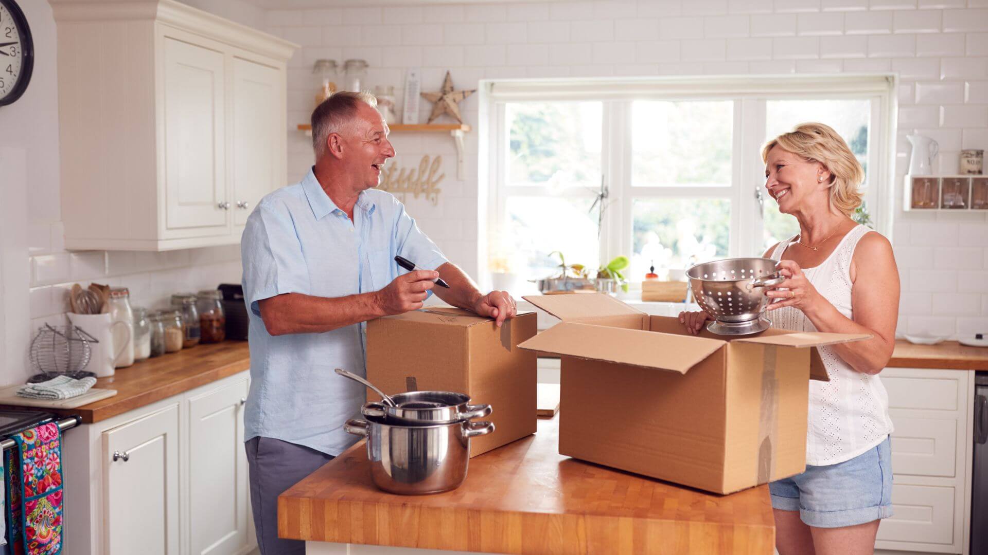 Downsizing your home. An older couple is happily packing kitchen items into boxes as they prepare to downsize their home, surrounded by a bright and inviting kitchen environment.