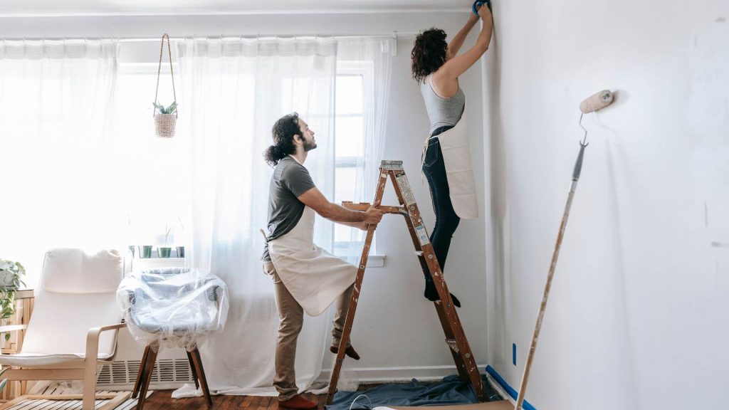 A couple working together to paint a wall in their home. They are standing on a ladder and making updates to their space, highlighting the decision of whether to renovate or sell their property.